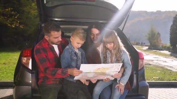 Вид спереди счастливых красивых родителей и их двух детей, которые собираются вместе, чтобы путешествовать на машине и с помощью карты найти правильный путь — стоковое видео