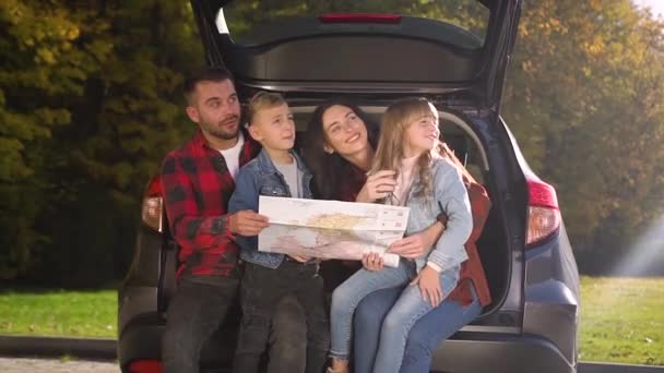 Atraktivní usměvavá rodina sedí v kufru auta, používá cestovní mapu a dívá se stranou, protože viděli něco zajímavého — Stock video