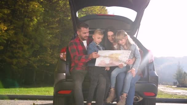 Feliz papá sonriente, mamá y dos niños sentados en el maletero y usando la hoja de ruta para encontrar el camino correcto para su viaje conjunto en un día soleado. — Vídeo de stock