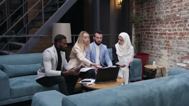 Успешные международные бизнесмены обсуждают новый проект и важные новые идеи, сидя на диване, используя цифровой компьютер в современном офисе, мозговой штурм — стоковое видео