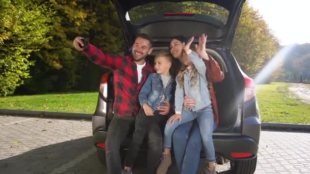 Привлекательный портрет радостной привлекательной семьи, которая сидит в багажнике и машет руками во время фотографирования на телефон отца — стоковое видео
