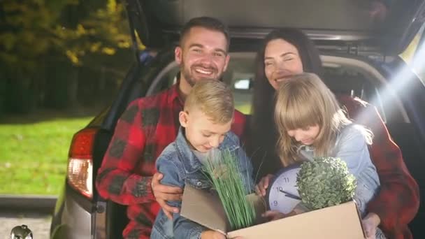 Felices padres guapos sosteniendo en sus rodillas lindos niños con caja de cartón con plantas antes de mudarse a su nueva casa — Vídeos de Stock