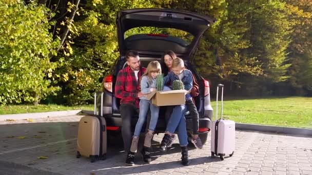 Aantrekkelijke trendy glimlachende ouders zitten in de kofferbak en houden op knieën kinderen met grote doos met bloemen tijdens hun verhuizing naar nieuwe flat — Stockvideo