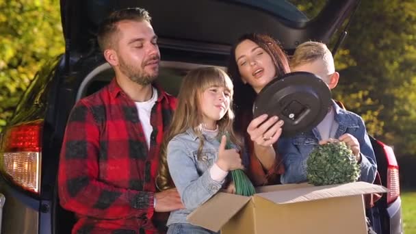 Felices niños sonrientes sosteniendo caja de cartón con plantas mientras están sentados en las rodillas de los padres y hablando de su reubicación en el nuevo apartamento — Vídeo de stock