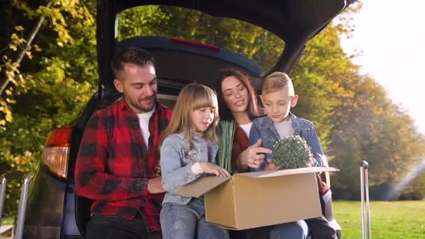 Sonriendo padres satisfechos con sus hijos agradables sentados en el maletero de autos y sosteniendo una caja grande con flores verdes durante su traslado a la nueva casa — Vídeo de stock