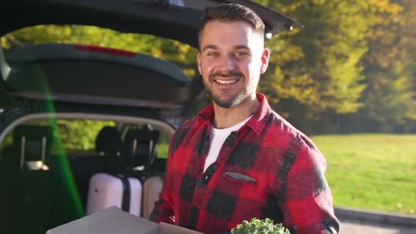 Bonito homem barbudo sorridente de 35 anos em roupas elegantes que estão perto do carro e segurando a caixa de papelão e olhando para a câmera — Vídeo de Stock