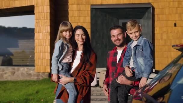 Vista cercana de la encantadora familia feliz joven que mira a la cámara con sonrisas lindas mientras está de pie cerca de la hermosa casa — Vídeo de stock