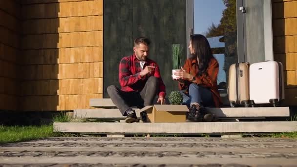 Bájos boldog fiatal pár szerelmes ül a lépcsőn a modern ház közelében, és tartja a kezükben zöld növények a dobozból — Stock videók