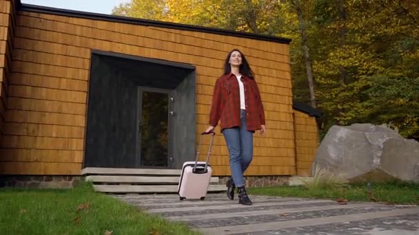 Satisfait jeune femme brune en vêtements décontractés portant valise sur roues à la voiture avant son voyage en voiture — Video