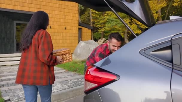 Vista lateral de la encantadora pareja joven y alegre de marido y mujer que descarga el maletero de los coches de cajas y maletas durante la mudanza a la nueva casa — Vídeos de Stock