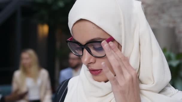 Close up portrait attractive muslim woman in glasses wearing white hijab headscarf looking at camera and smiling happily standing on the office colleagues background — Stock Video