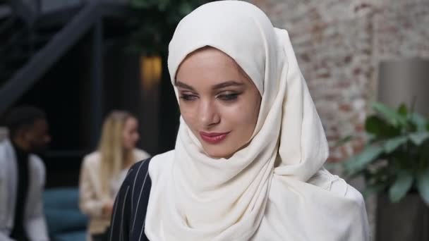 Close-up de rosto retrato de mulher muçulmana atraente em hijab branco lenço de cabeça olhando para a câmera e sorrindo de pé no escritório colegas fundo — Vídeo de Stock