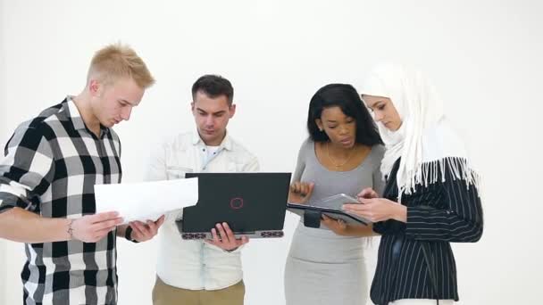 Уверенные молодые два европейских мужчины и две африканские американские и мусульманские женщины, использующие ноутбук, планшет ПК, отчеты и различные документы во время работы в рамках совместного проекта — стоковое видео