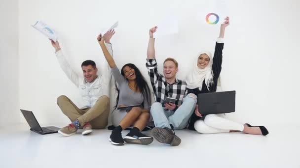 Happy smiling mixed race office coworkers sitting near the wall and waving with raised hands after successful ending joint work — Stockvideo