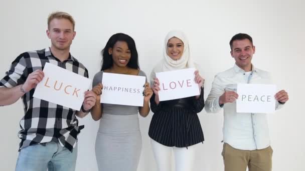 Attrayant joyeux 30s hommes et femmes multiethniques debout près du mur blanc et tenant dans les mains des affiches avec des mots écrits amour, paix, chance et bonheur — Video