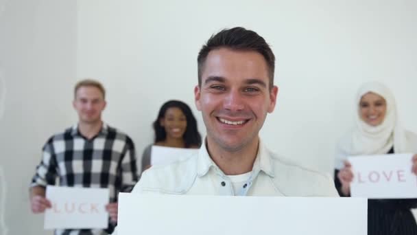 Sonriente joven alegre posando en cámara con cartel con palabras "No racismo" en el fondo de sus compañeros multiétnicos con inscripciones "Love.Luck.Happiness " — Vídeos de Stock