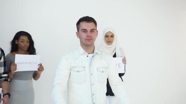 Ernster attraktiver junger Mann mit Plakat "kein Rassismus", der auf den Hintergrund seiner multirassisch lächelnden Freunde mit der Aufschrift "luck.happiness.love" — Stockvideo