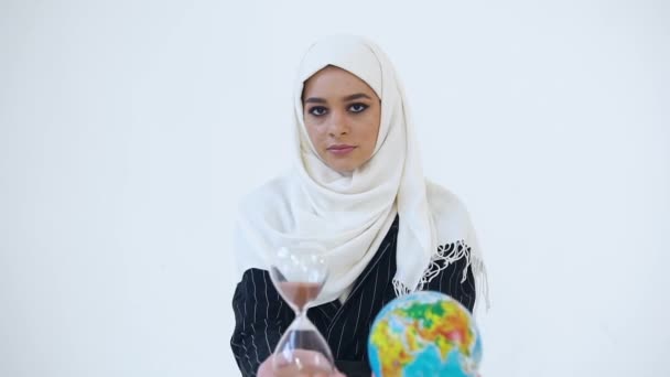 Привлекательная молодая серьезная арабская леди в хиджабе держит в руках песочные часы и маленький глобус глядя на камеру на белом фоне — стоковое видео