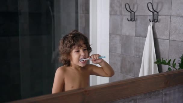Красивый маленький мальчик с каштановыми вьющимися волосами пользуется зубной пастой и зубной щеткой, когда чистит зубы перед или после сна перед зеркалом в ванной комнате — стоковое видео