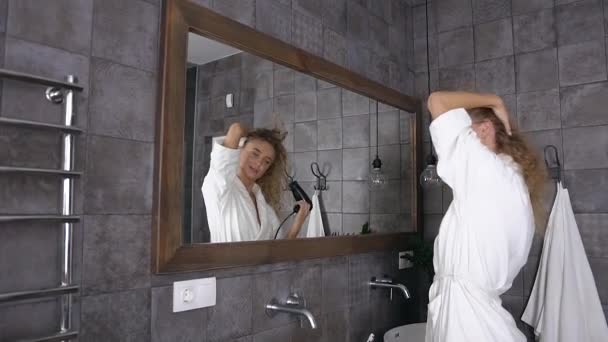 Attractive blonde woman in white bathrobe with long curly hair drying hair in front of mirror in bathroom at home — 비디오