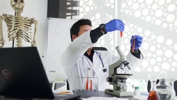 Młody skoncentrowany azjatycki lekarz w białej sukni i niebieskich rękawiczkach porównujący wypełnienie dwóch probówek czerwonymi roztworami i zapisanie znaków na papierze i komputerze — Wideo stockowe