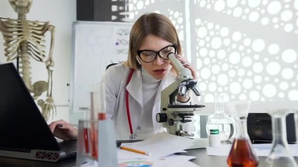 Gözlüklü hoş görünümlü genç doktor klinik dolabında mikroskop ve bilgisayarla çalışıyor. — Stok video