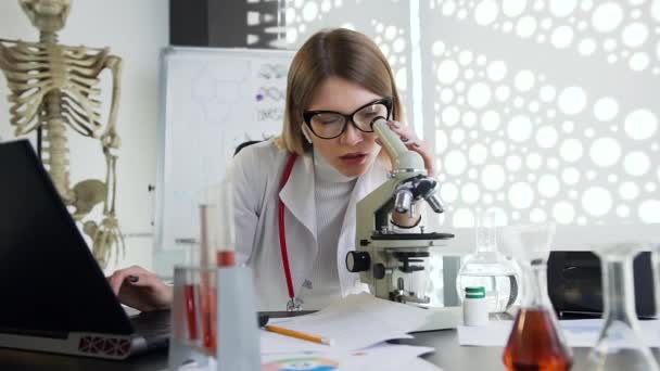 Buen aspecto médico joven agradable en gafas que trabajan con microscopio y computadora en el gabinete de la clínica — Vídeo de stock