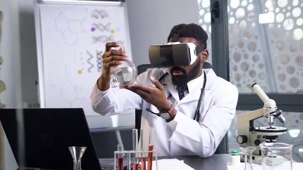 Целеустремленный 35-летний бородатый африканский врач-мужчина смотрит на фляжку с химической жидкостью с помощью гарнитуры виртуальной реальности — стоковое видео