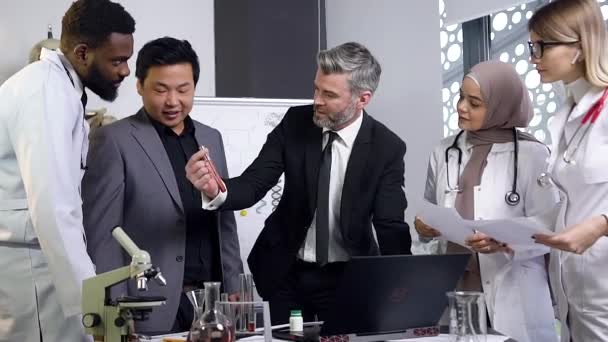 Good-looking respectable gray-haired male head doctor telling to his mixed race doctors-colleagues about chemical properties liquids — 비디오