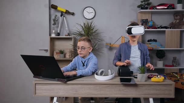 Atractivo retrato de dos chicos que pasan tiempo en la habitación de los niños, uno de ellos jugando en el ordenador, otro en auriculares de realidad virtual — Vídeos de Stock