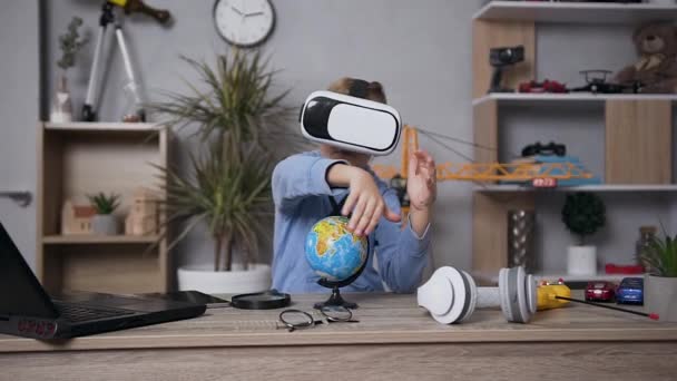 Attractive concentrated 6-aged boy sitting at the table in his room and studing the structure of the globe using special virtual 3d glasses — Stock Video