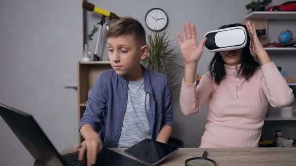 Прекрасна усміхнена брюнетка жінка в гарнітурі віртуальної реальності сидить за столом і веселий хлопчик-підліток стоїть поруч і використовує ноутбук — стокове відео