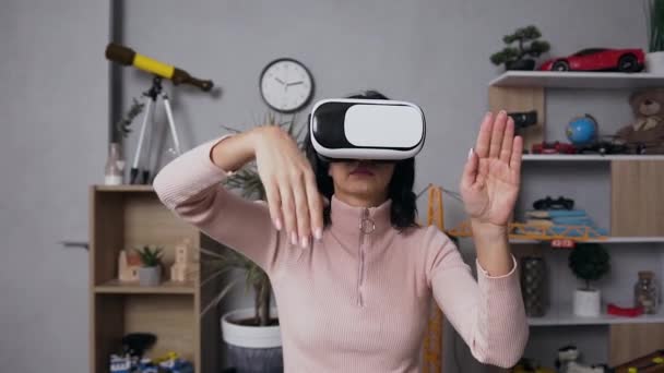 Belle jeune femme concentrée en vêtements décontractés assis sur le lieu de travail à la maison et faisant différents mouvements des mains dans l'air à l'aide de lunettes 3D virtuelles — Video