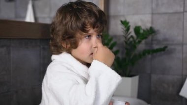 Siyah kıvırcık saçlı, beyaz cüppeli yakışıklı çocuk banyoda duş aldıktan sonra burnuna krem sürüyor..