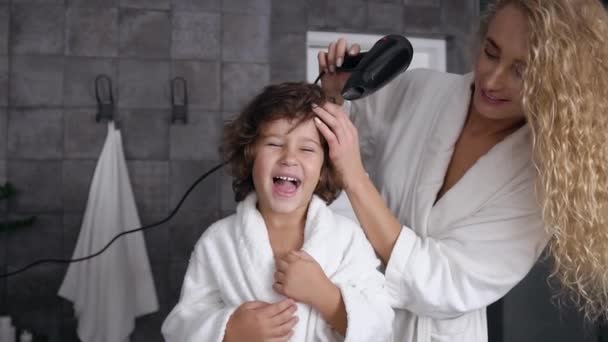 Caring young mom with long curly hair dries curly hair with a hairdryer her little happy son after taking shower at bathroom — Stock Video