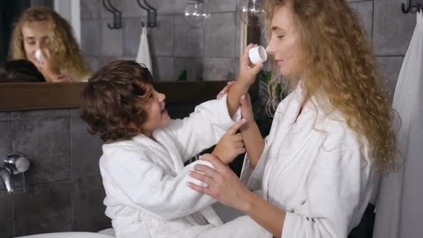 Der kleine lächelnde Junge trägt Hautpflegecreme auf sein schönes Gesicht auf. Familien-, Hygiene- und Gesundheitskonzept — Stockvideo