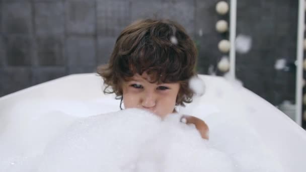 Портретное лицо маленького милого мальчика с темными кудрявыми волосами, играющего с водой и пеной во время купания в ванне — стоковое видео