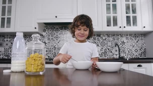 Feliz niño pequeño sentado en la mesa de la cocina y come con placer desayuno saludable de copos de maíz y leche en la cocina casera — Vídeos de Stock