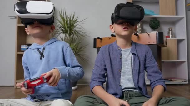 Chicos adolescentes modernos de buen aspecto sentados en la alfombra y jugando juegos con auriculares de realidad virtual y joysticks — Vídeo de stock