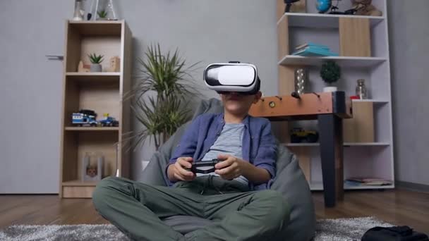 Attractive portrait of joyful smart 12-aged boy which playing video game on the bean bag sofa using virtual reality headset and remote controller — Stock Video