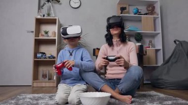 İyi görünümlü, modern anne ve oğul halının üzerinde oturmuş video oyunu oynarken özel sanal 3D gözlükler takıyorlar.