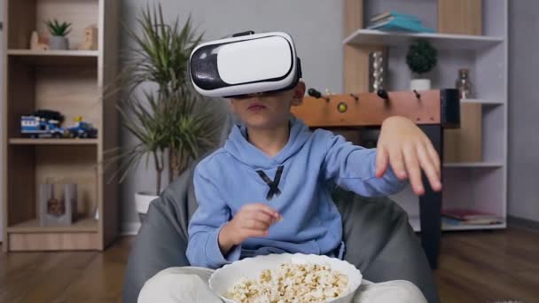 Vista frontal de belo focado menino de 10 anos em realidade virtual fone de ouvido que sentado no sofá confortável saco de feijão, comer pipocas e desfrutar de vídeo — Vídeo de Stock