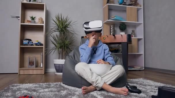 Vista frontal de niño guapo concentrado en auriculares de realidad virtual sentado en un sillón inflable disfrutando de video y comiendo palomitas de maíz — Vídeos de Stock