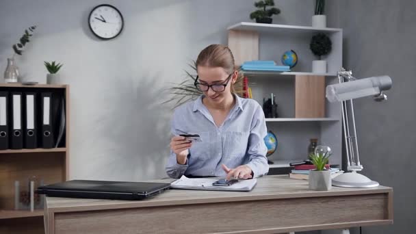 Добродушная счастливая молодая женщина сидит за столом и кладет данные кредитной карты на телефон, чтобы сделать онлайн покупки — стоковое видео