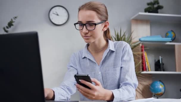 Attraktivt porträtt av snygg glad leende kvinna i glasögon som sitter i moderna skåp och arbetar med hjälp av datorn och hennes telefon — Stockvideo