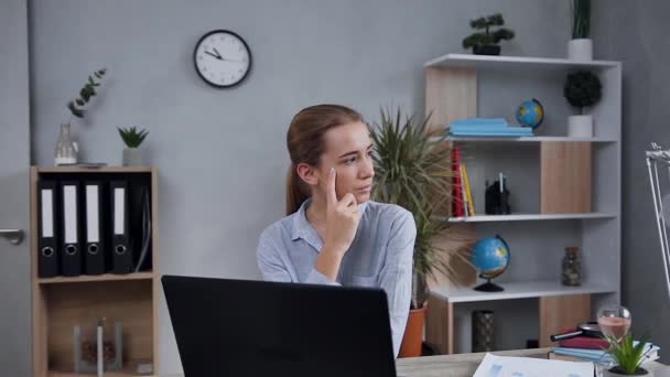 Gut aussehende junge blonde Dame in lässiger Kleidung, die am Tisch sitzt, während sie am Computer arbeitet und wegschaut — Stockvideo
