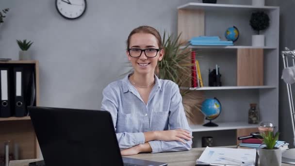 Portrait attrayant de souriante élégante femme de 25 ans dans des lunettes qui posent sur la caméra avec un sourire sincère à son lieu de travail avec ordinateur — Video