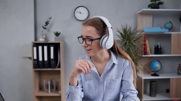 Close up of pretty satisfied blond woman in earphones which having videocall with somebody while writing down important notes — Stock Video