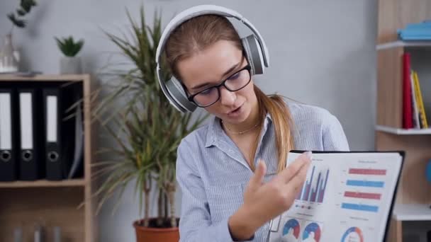 Mujer joven satisfecha agradable de gran espíritu en auriculares que tienen videocall hablando con el cliente y discutiendo datos de informes — Vídeos de Stock