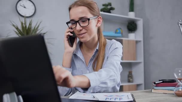 Καλοδιάθετη χαμογελαστή σύγχρονη 25χρονη κοπέλα με γυαλιά που μιλάει στο τηλέφωνο κατά τη διάρκεια της εργασίας στον υπολογιστή και κρατώντας σημειώσεις στην έκθεση — Αρχείο Βίντεο